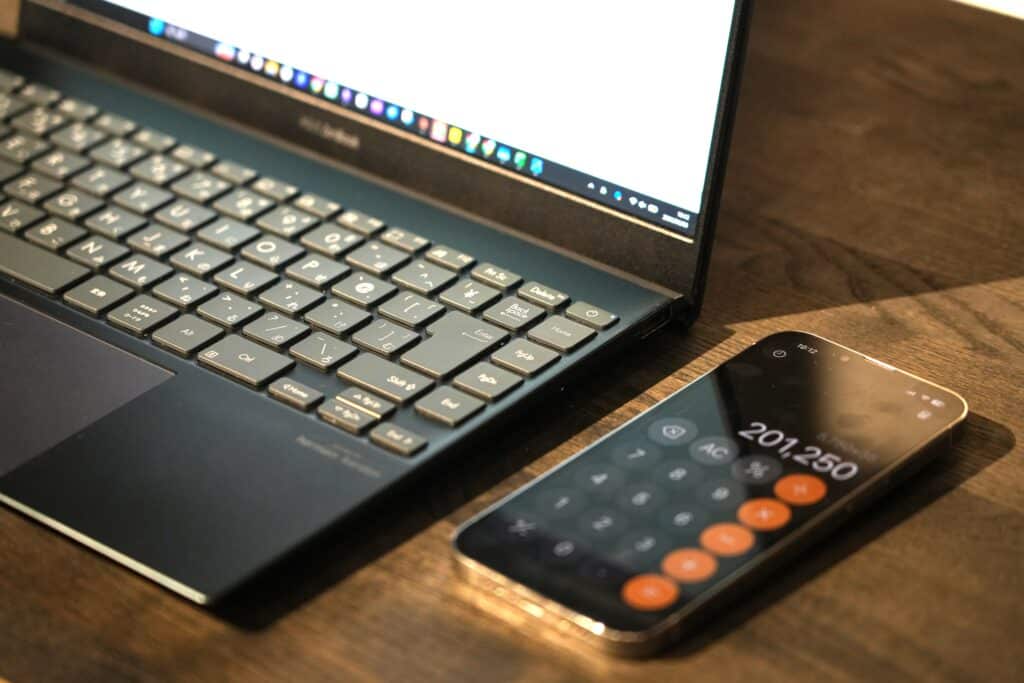 木の机の上に、ノートパソコンと、201,250と表示された電卓アプリが起動したスマートフォンが置かれている