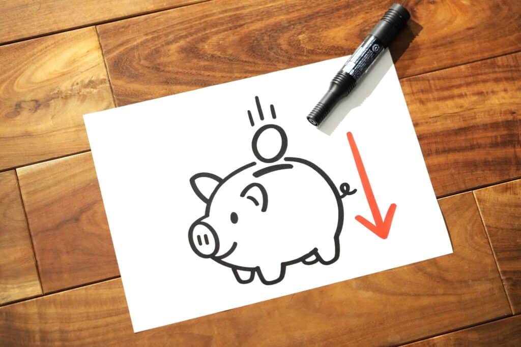 白い紙の上に、貯金箱と硬貨、そして下向きの赤い矢印が描かれており、その上に黒いマーカーが置かれている