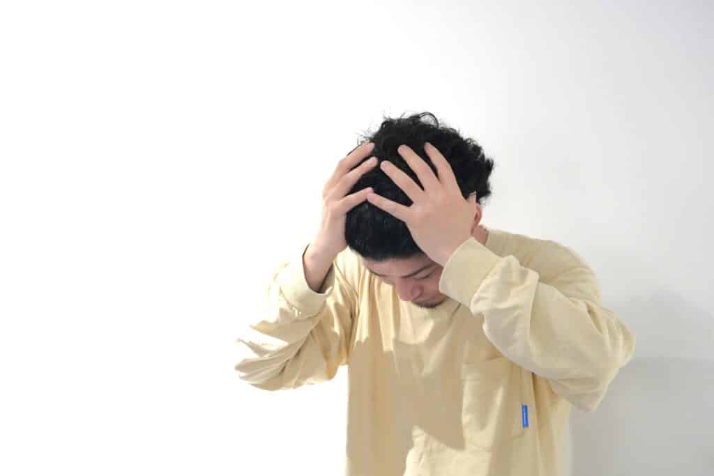 白い背景の前で、ベージュの長袖を着た人が頭を抱えてうつむいている