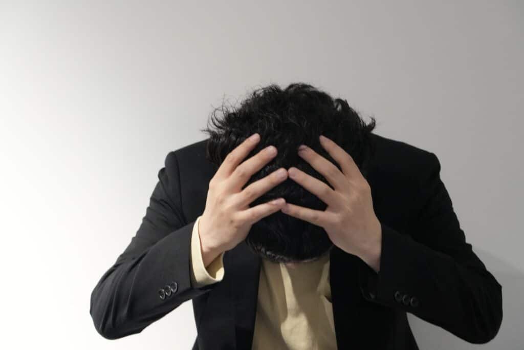 黒いブレザーの人が頭を抱えて座っている薄い背景.