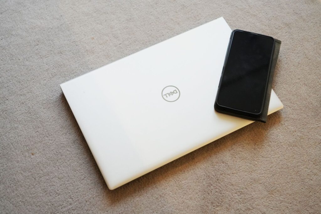白いDellのノートパソコンの上に黒いスマートフォンが置かれており、床はベージュのカーペットだ