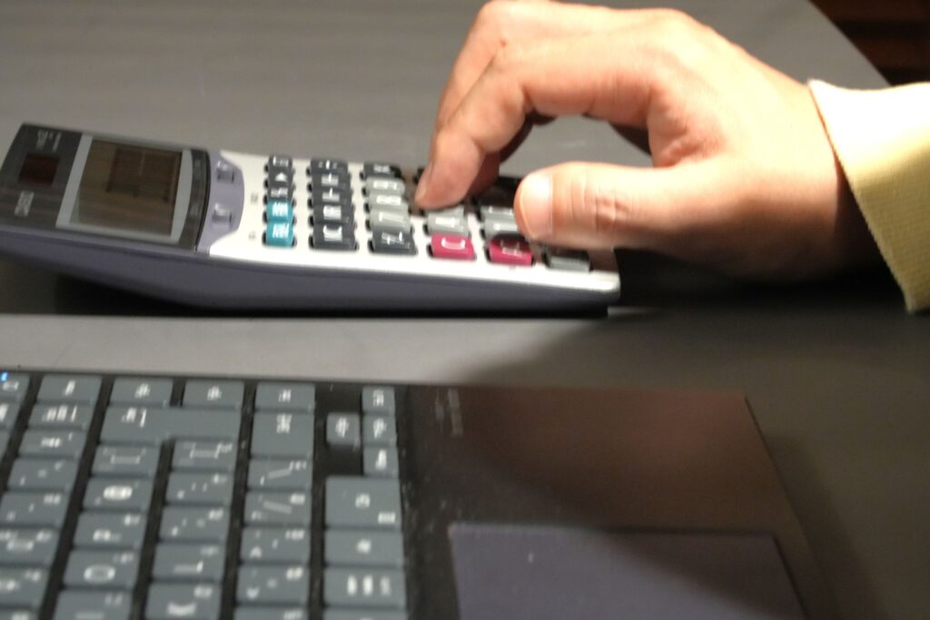 手が電卓のボタンを押していて、その隣にはノートパソコンのキーボードがある