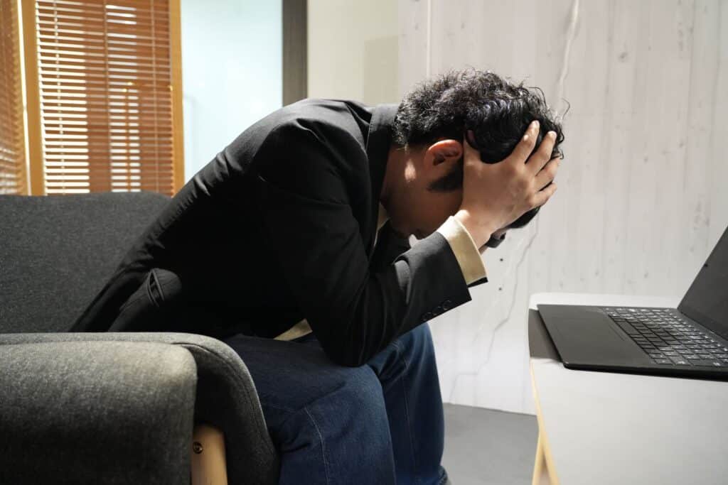 スーツ姿の男性がソファに座り、頭を抱えている。その前のテーブルにはノートパソコンが置かれている