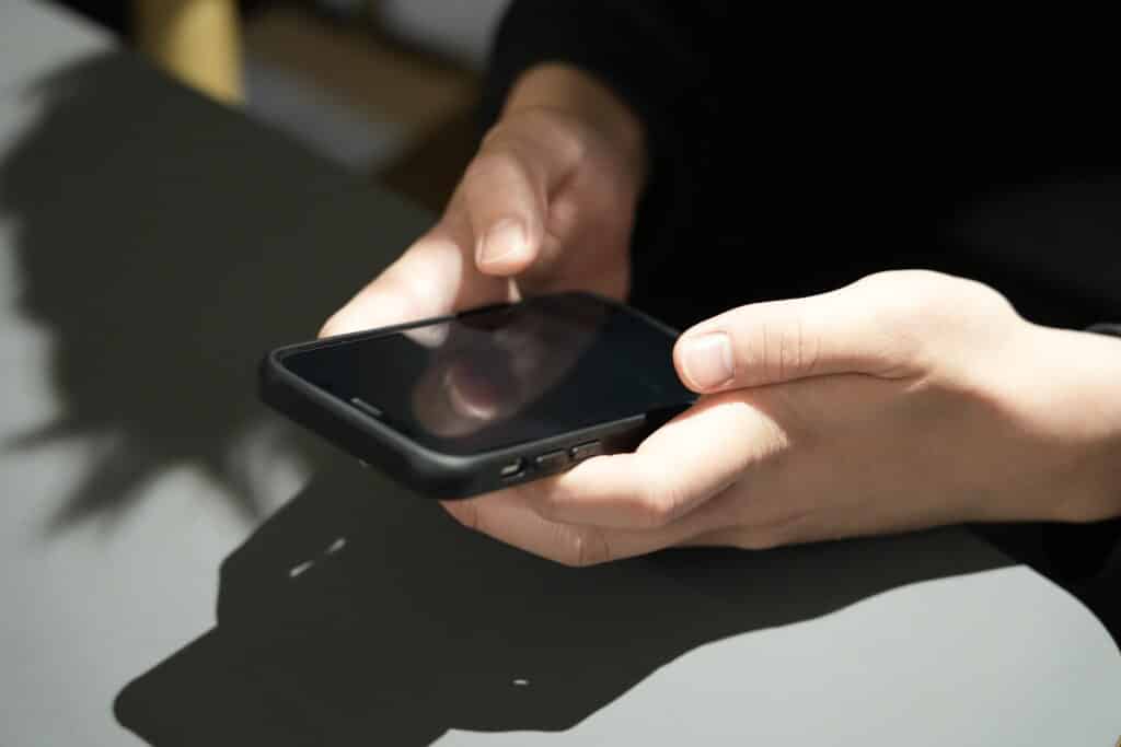 背景には、黒いケースのスマートフォンを手に持ち、操作している人物の灰色の顔が映っている