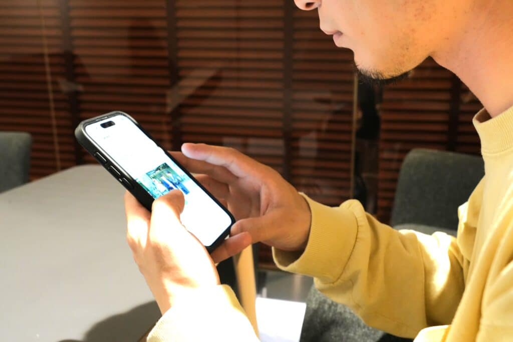 黄色い服を着た人がテーブルで両手でスマートフォンを操作している背後に、木製のブラインドがある